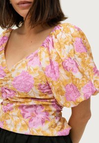 Blumenbluse mit kurzen Puffärmeln. Mit einem rosa und gelben Blumenmuster auf einem hellen Hintergrund. Weicher Stoff, V-Ausschnitt-Design.