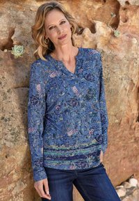 Langärmliges blaues Oberteil mit floralem und Paisleymuster, tiefem V-Ausschnitt und weichem, strukturiertem Stoff. Getragen mit dunklen Jeans.