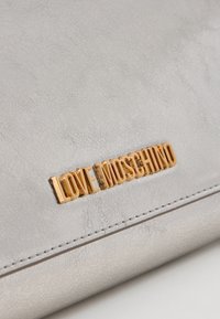 Superficie in pelle grigia chiara con texture, con lettering in metallo dorato che scrive "LOVE MOSCHINO" sopra una cucitura.