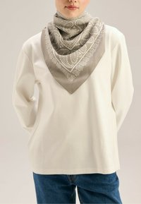 Jedwabny szal ma szary wzór paisley z białymi akcentami, w kształcie trójkąta, noszony na szyi na białej bluzce z długim rękawem.