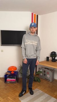 Pelēks džemperis ar grafisko dizainu, tumši zili svārki un melni sporta apavi ar baltiem svītrām. Iekļauta arī zila cepure. Iekštelpu vide ar augiem.