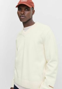 Young man wearing a cream V-neck sweater, light shirt underneath, rust-orange cap, and small hoop earring, standing against a plain white background.