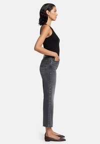 Schwarzes Tanktop und graue, hochgeschnittene Jeans mit schmaler Passform, die rohe Säume haben. Getragen mit braunen Ballerinas. Seitenansicht.