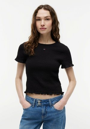 Kobieta z falującymi brązowymi włosami, ubrana w dopasowaną, czarną, teksturowaną bluzkę z krótkim rękawem i wysokim stanem niebieskie jeansy, ręce w kieszeniach, neutralne tło.