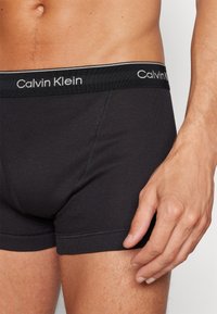Schwarze Baumwoll-Boxershorts mit einer angepassten Passform, ausgestattet mit einem Calvin Klein Logo-Bund und sichtbaren Nähten. Eine Hand ruht auf dem Stoff.