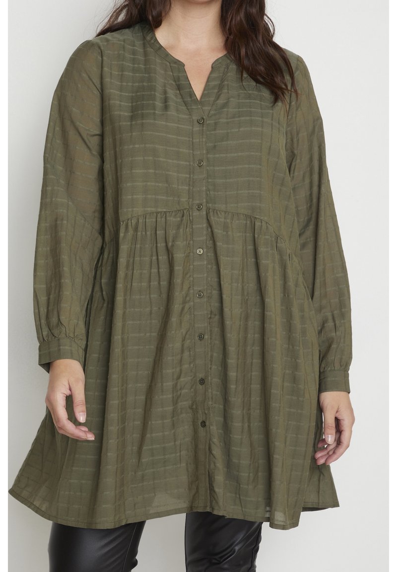 Femme portant une robe olive fluide boutonnée avec de subtiles rayures horizontales et des manches longues sur un pantalon noir.