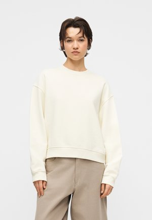 Junge Person mit kurzen dunklen Haaren, die ein lockeres cremefarbenes Sweatshirt und beige Hosen mit weitem Bein trägt, vor einem einfachen hellen Hintergrund stehend.