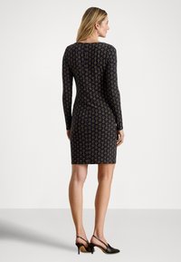 Svart langermet kjole med et gjentakende gull og hvitt mønster. Slank passform med midt-lårlengde, kombinert med svarte høyhælte sko.