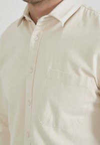 Gros plan sur une personne portant une chemise à boutons de couleur crème avec une poche sur la poitrine et un col légèrement ouvert.