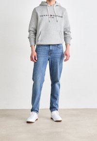 Felpa grigia chiara con tasca frontale, con logo rosso e blu. Abbinata a jeans azzurri e sneakers bianche con suole marroni.
