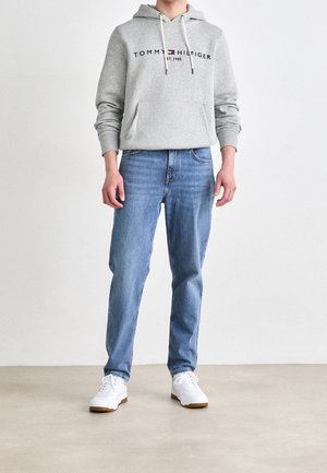 Felpa grigia chiara con tasca frontale, con logo rosso e blu. Abbinata a jeans azzurri e sneakers bianche con suole marroni.