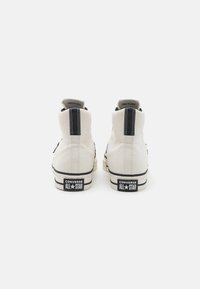 Adidași înalți din pânză albă cu accente negre, având toc întărit, cusături contrastante și un logo vizibil pe spate.