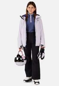 Ljuslila jacka med kontrasterande blå dragkedja, bärs över en marinblå fleece, i kombination med svarta byxor. Håller en vit hjälm och handskar.