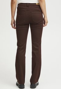 Jean évasé marron avec une texture lisse, cinq poches et une étiquette à la taille. Le pantalon a une coupe ajustée et est de longueur cheville.