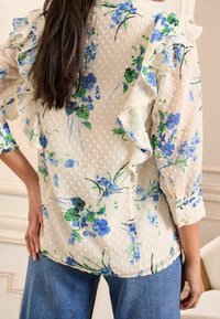 Durchsichtige Blumenbluse mit blauen und grünen Mustern auf einem cremefarbenen Hintergrund, mit gerafften Ärmeln und einem strukturierten Punkte-Design.