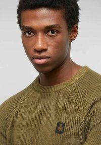 Giovane uomo con capelli corti e ricci, che indossa un maglione a maglia testurizzata di colore verde oliva con una piccola toppa logo nera e arancione.