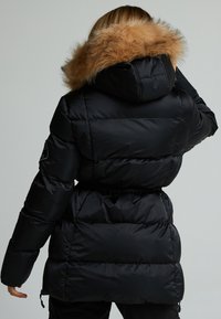 Giacca a vento nera con vita aderente, dotata di cappuccio staccabile in pelliccia sintetica color beige. Il tessuto appare lucido con sezioni trapuntate.