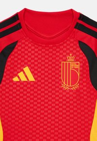 Κόκκινη αθλητική φανέλα με μαύρες και κίτρινες λεπτομέρειες, που διαθέτει υφή και λαιμόκοψη, με το έμβλημα της Βασιλικής Ομοσπονδίας Ποδοσφαίρου του Βελγίου προεξέχον.