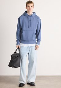 Giovane uomo che indossa un hoodie blu, jeans larghi di lavaggio chiaro, scarpe nere, tiene in mano una borsa in pelle nera, in piedi contro un muro semplice.