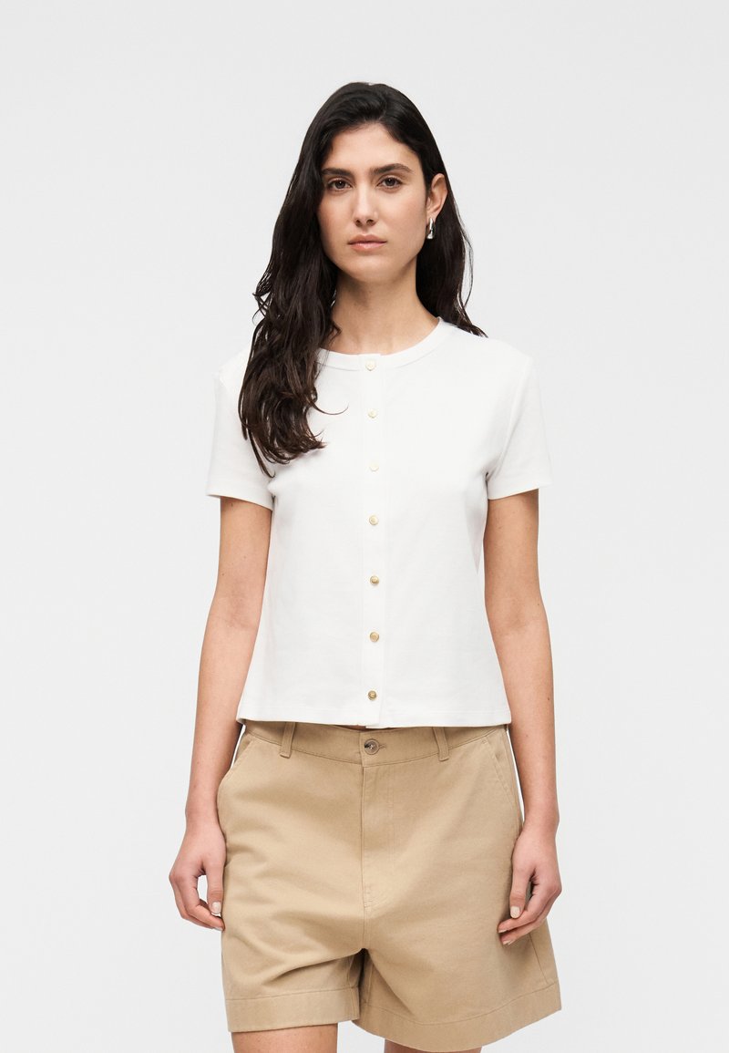 Λευκό πουκάμισο με κοντά μανίκια και στρογγυλή λαιμόκοψη, με σειρά από κουμπιά σε χρυσή απόχρωση στο μπροστινό μέρος. Φορεμένο με μπεζ σορτς.