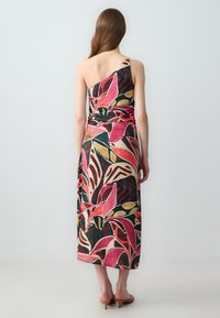 Frau mit langen braunen Haaren, die ein bunten, floralen Midikleid mit einem Schulterträger und beigefarbene High Heels trägt, steht vor einem schlichten, hellen Hintergrund.