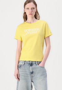 Gul bomull T-skjorte med korte ermer, med "tommy jeans" logo i hvitt. Kombinert med lyse, avslappede denimshorts.