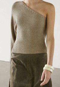 Pull beige tricoté avec une épaule découverte, tissu texturé. Associé à une jupe en daim marron et de larges bracelets crème.