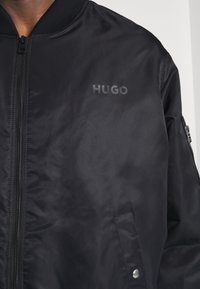 Giacca bomber nera con colletto a costine, chiusura con zip e tasca laterale. Presenta un logo tonale "HUGO" sul petto. Tessuto liscio e leggero.