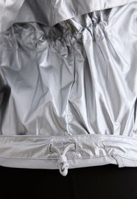 Veste argentée réfléchissante avec détail élastique froncé et cordon blanc ajustable à la taille sur pantalon noir.