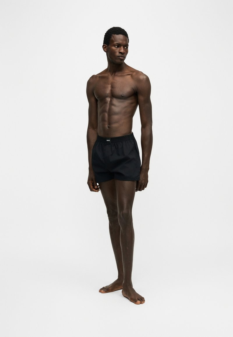 Homme mince debout pieds nus portant un boxer noir, regardant sur le côté sur un fond clair uni.