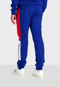 Pantaloni tuta blu con una striscia laterale rossa, caratterizzati da testo bianco "LE COQ SPORTIF" su una gamba. Realizzati in tessuto morbido, con una vestibilità comoda.