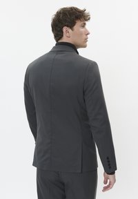 Hallituspruun ülikonna jakk, millel on struktureeritud disain, tekstureeritud kangas, nurgakrae ja neli nupuvajutust varrukatel. Tagantvaade on välja toodud.