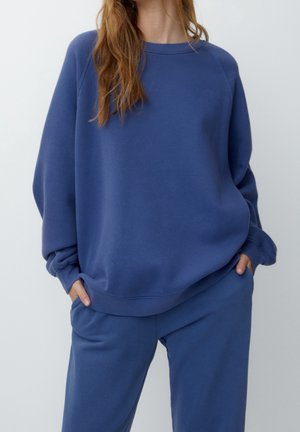 Personne portant un sweat-shirt bleu ample et un pantalon assorti, les mains dans les poches, sur un fond uni.