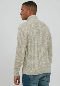 Crème gebreide trui met een hoge kraag, voorzien van gestructureerde patronen en geribde manchetten. Gecombineerd met blauwe jeans, wat een casual stijl benadrukt.