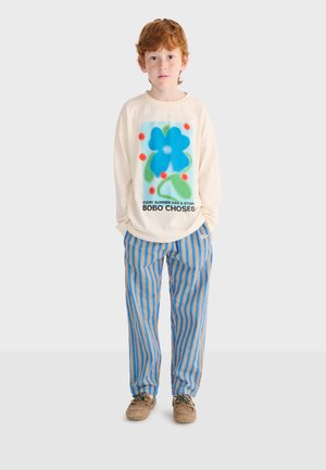 Ragazzo dai capelli rossi che indossa una maglia a maniche lunghe color crema con una grafica di un fiore blu e pantaloni a righe blu e arancioni, in piedi su uno sfondo bianco.