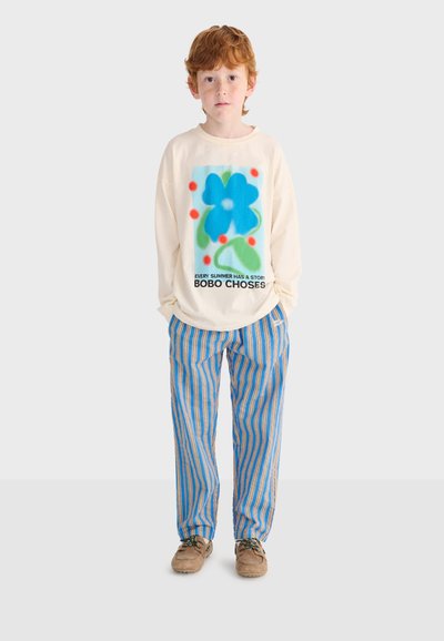 Red-haired boy wearing a cream long-sleeve shirt with a blue flower graphic and striped blue and orange pants, standing against a white background.