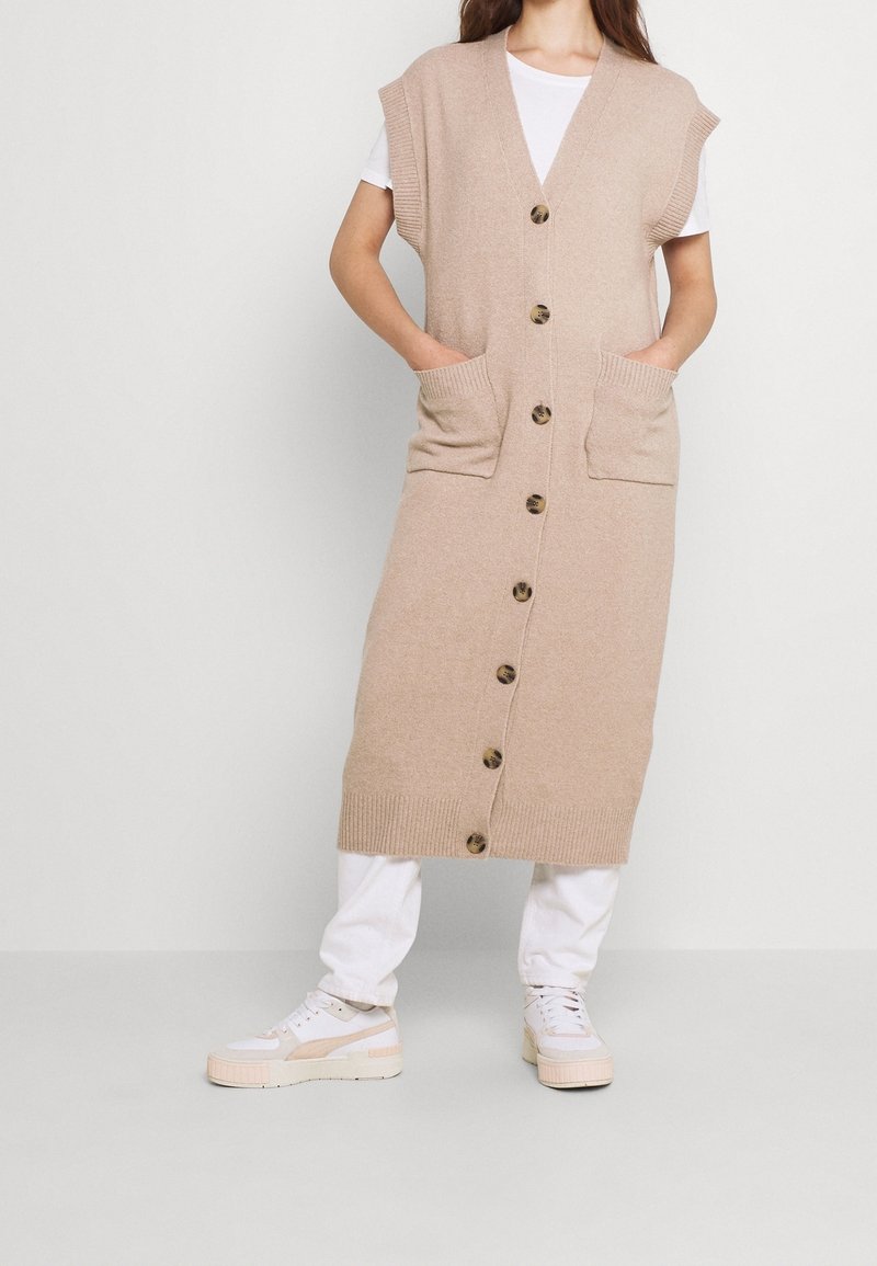 Béžová dlouhá pletená vesta s krátkými rukávy, zapínáním vpředu na knoflíky a dvěma kapsami. Nošena přes bílé tričko a doplněna bílými sportovními botami.