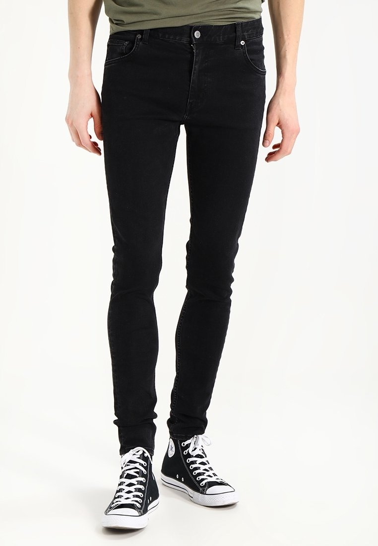 Personne portant un jean slim noir et des baskets montantes noires avec des lacets blancs, debout devant un fond blanc.