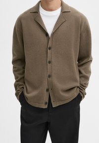 Homme portant un cardigan marron boutonné sur une chemise blanche, les mains dans les poches, associé à un pantalon noir, sur un fond clair uni.