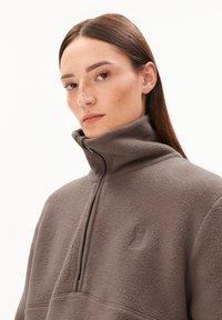 Pullover in pile di colore marrone tenue, con colletto alto e chiusura a zip a quarti. Dettaglio del logo ben visibile sul petto, texture morbida.