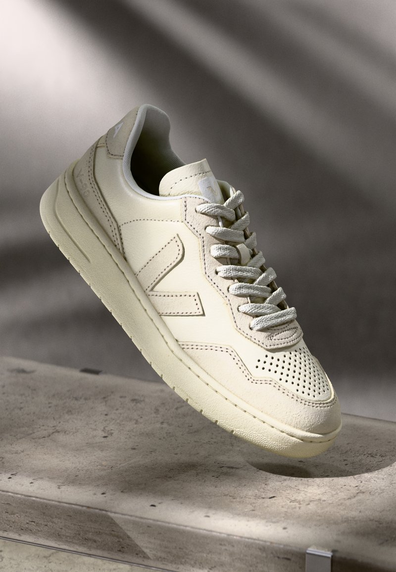 Baskets en cuir beige avec embout perforé, accents texturés, lacets plats et semelle épaisse, arborant un logo visible sur le côté.