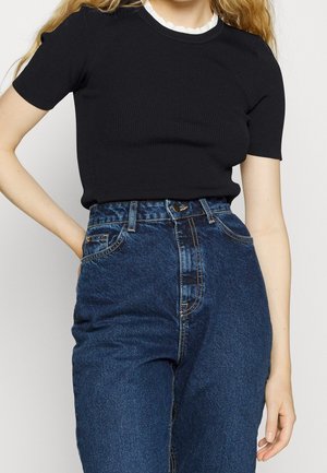 Frau trägt ein schwarzes, kurzärmeliges Strickoberteil und hochtaillierte dunkelblaue Jeans, stehend vor einem schlichten Hintergrund.