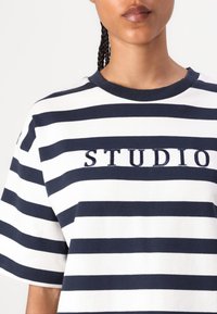 Camisa de mangas cortas a rayas en algodón azul marino y blanco, con el logotipo bordado "STUDIO" en el pecho y un cuello redondo clásico.