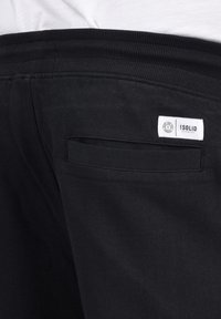 Pantalons de survêtement noirs avec une taille côtelée, présentant une petite étiquette et un détail de poche arrière. Le tissu semble lisse et doux.