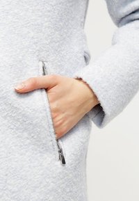 Cappotto esterno azzurro chiaro, con texture e tasca zip laterale. Tessuto morbido, vestibilità su misura, con finiture pulite sui polsini.