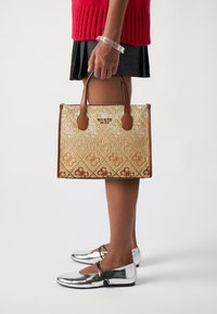 Tan and gold tote bag with a patterned exterior, brown handles, and a structured rectangular shape, paired with silver flat shoes.