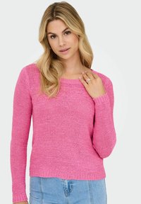Une femme blonde aux ondulations lâches porte un pull en tricot rose vif et un jean bleu clair, touchant une bague argentée sur sa main près de son épaule.