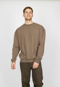 Sudadera marrón con escote redondeado y hombros caídos, que presenta mangas largas y un dobladillo acanalado, combinada con pantalones oscuros.