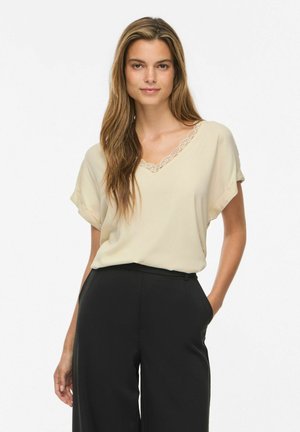 Kvinde med langt hår iført en beige kortærmet bluse med blonderkant og sorte højtaljede bukser, stående med den ene hånd i lommen.