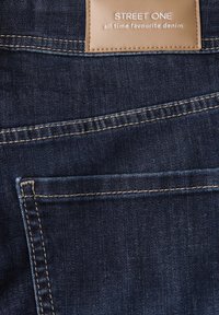 Tessuto in denim blu scuro con texture visibile, dotato di un'etichetta in pelle beige con la scritta "STREET ONE" e dettagli di cucitura. Include una tasca posteriore.